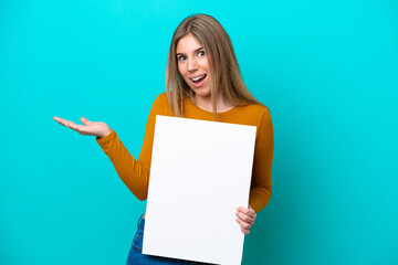 Young caucasian woman isolated on blue background holding an empty placard with surprised expression