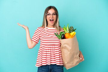 Young woman holding a grocery shopping bag isolated on blue background with shocked facial...