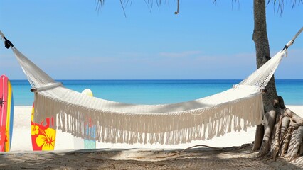 White hammock tied between palm trees on tropical beach with clear turquoise water, surfboards...