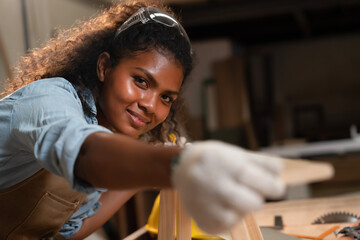 African American young carpenter woman with smiling face and safety glasses overhead build small chairs, using tape measure wooden planks before making a chair assembly in wooden factory