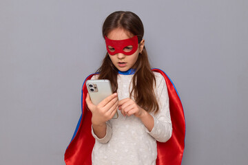 Shocked sad little girl wearing superhero costume and mask isolated over gray background using cell phone for online communicating reading message with bad news