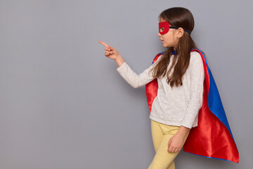Surprised little girl wearing superhero costume and mask isolated over gray background indicating...
