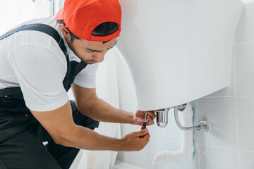 Skilled plumber using a wrench repairs a water pipe under a bathroom sink. His focus is on...