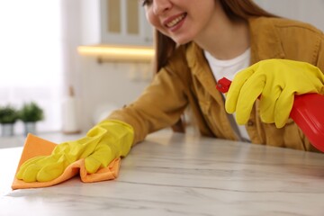 Woman with spray bottle and microfiber cloth cleaning white marble table in kitchen, closeup