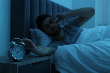 Man suffering from headache and turning off alarm clock in bed at night, selective focus