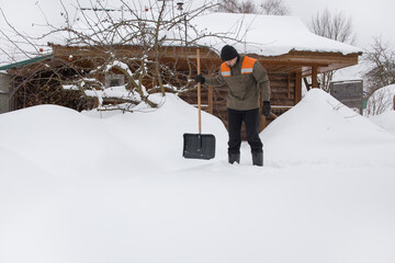 A man in work clothes stands with a shovel in his hands and looks at the fallen snow.