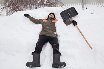 A man in work clothes lies in a snowdrift and holds a shovel in his hand.