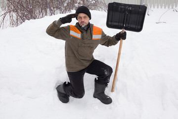A funny man with a shovel in his hands stands near a snowdrift and shows a hand gesture.