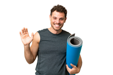 Young sport man going to yoga classes while holding a mat over isolated chroma key background saluting with hand with happy expression