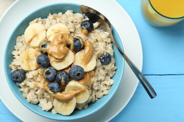 Tasty oatmeal with banana, blueberries and peanut butter served in bowl on light blue wooden table, top view