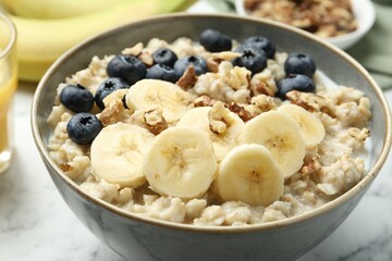 Tasty oatmeal with banana, blueberries, walnuts and milk served in bowl on white marble table, closeup