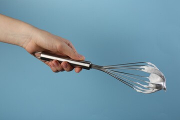 Woman holding whisk with whipped cream on light blue background, closeup
