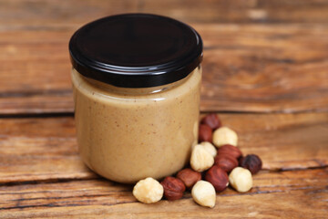 Tasty nut paste in jar and hazelnuts on wooden table, closeup