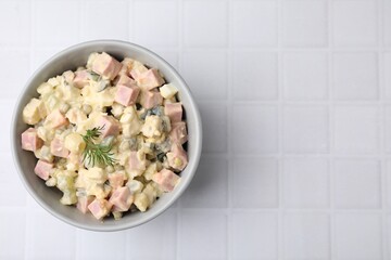 Tasty Olivier salad with boiled sausage in bowl on white tiled table, top view. Space for text