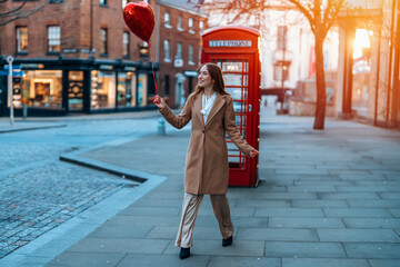 young happy woman with a heart-shaped balloon falling a love, having a fun day, walking against the red phone box in English city Spring is in the air Lifestyle, tourism, valentines day concept - obrazy, fototapety, plakaty