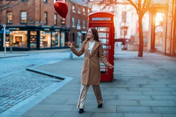 Fotobehang young happy woman with a heart-shaped balloon falling a love, having a fun day, walking against the red phone box in English city Spring is in the air Lifestyle, tourism, valentines day concept © Iryna
