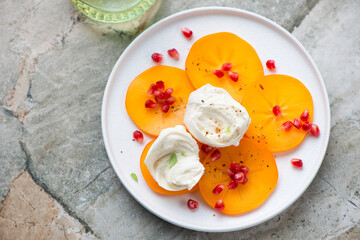 Persimmon slices with mozzarella and pomegranate seeds on a white plate, horizontal shot on a grey granite background, high angle view
