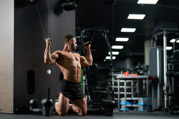 Fototapeta na wymiar Strong sportsman kneeling, while doing exercise for biceps in gym. Full body of muscular male athlete wearing black shorts, pulling cables of training apparatus indoors. Concept of sport, workout.