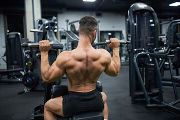Fototapeta na wymiar Muscular male bodybuilder exercising with training apparatus in gym. Back view of anonymous strong shirtless man, building back muscles, working out at evening. Sport, lifestyle, bodybuilding concept.
