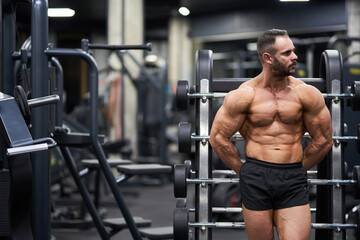 Pensive shirtless crossfit athlete leaning on palf rack barbell platform, having rest after training. Front view of strong, muscular guy looking away, while exercising in gym. Concept of sport.
