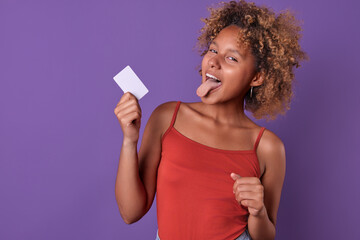 Young cheerful pretty African American woman teen holds credit card in hands to make online purchases and use cashless payments when traveling around world stands on purple background.