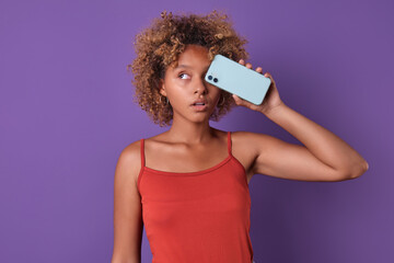 Young beautiful mysterious African American woman student puts mobile phone to eye and looks to side advertising new model of popular smartphone dressed in casual style stands on purple background.