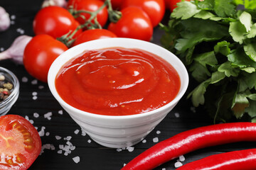 Delicious ketchup in bowl, products and salt on black wooden table, closeup. Tomato sauce