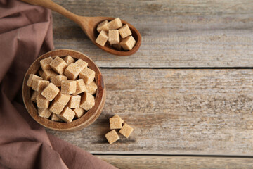 Bowl and spoon with brown sugar cubes on wooden table, flat lay. Space for text