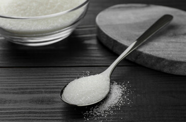 Spoon with granulated sugar on black wooden table, closeup