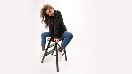 Charming caucasian woman in blue jeans and black jacket sitting on small wooden ladder over grey...