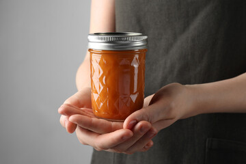 Woman holding glass jar of delicious persimmon jam on gray background, closeup. Space for text - Powered by Adobe