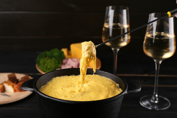 Dipping piece of ham into fondue pot with melted cheese on table, closeup