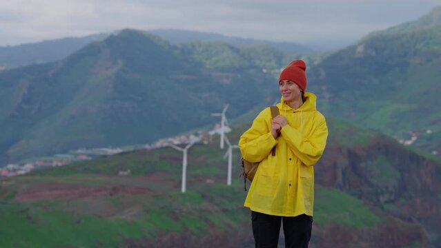 Attractive traveling woman in a yellow jacket and backpack looking at windmills in field 