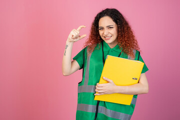 Fototapeta premium Front view of pretty, young female wearing green vest, looking at camera, squint, raising hand, showing. Isolated on pink studio background.