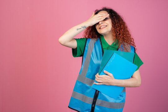 Front view of pertty, female paramedic wearing vest, standing, raising hand, closing eyes, smiling. Isolated on pink studio background.