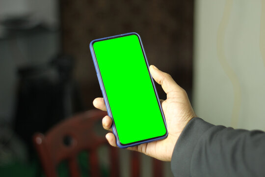 Hand holding smartphone with green screen on blurred background for easy replacement.