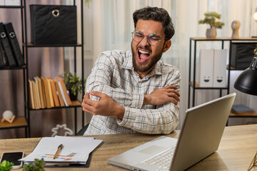 Fototapeta premium Tired Indian Arabian businessman guy manager sitting at home office table with laptop and experiencing pain and discomfort in elbow joint and massaging painful hand. Arthritis and bursitis concepts