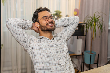 Happy overjoyed Indian businessman working on laptop at home office desk putting on earbuds listening to favorite energetic disco music. Hispanic guy relaxing, taking a break with hands behind head.