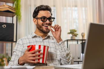 Indian young businessman freelancer taking break from work wearing 3D glasses earing popcorn and watching movie on laptop. Smiling Hispanic man sitting at home office desk having snacks during break.