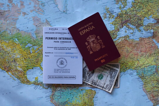 Girona, España. September 8th 2023. International Driver's License and Spanish passport for travelling around the world. With some dollars and an atlas mapamundi background.