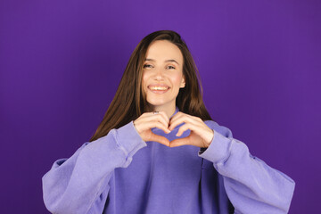 Young brunette woman wearing casual clothes smiling in love doing heart symbol shape with hands isolated on purple background. Romantic concept.