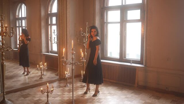 A woman wearing a black dress stands in front of a mirror in a room with candles