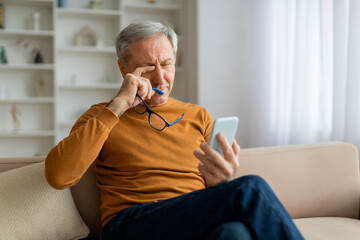 Tired senior man rubbing his eyes, using smartphone at home