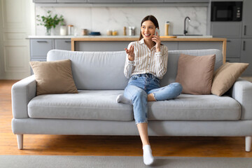 Cheerful young woman chatting on cellphone, sitting on sofa