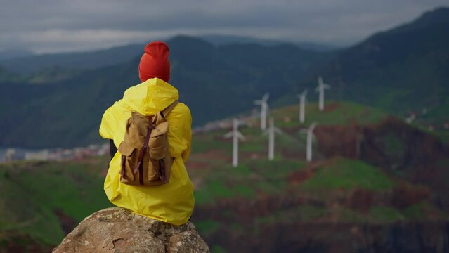 Attractive traveling woman in a yellow jacket and backpack looking at windmills in field 