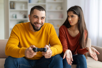 Angry young woman looking at boyfriend playing video games at home