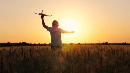 Happy male teenager running with aircraft toy flying pilot imagination at sunset sunrise wheat field back view. Joyful boy kid child silhouette playing plane plaything fantasy flight game meadow dusk