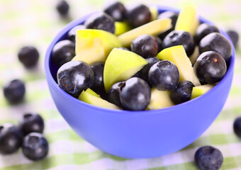 A blue bowl full of blueberries and pieces of apple with blueberries around it.