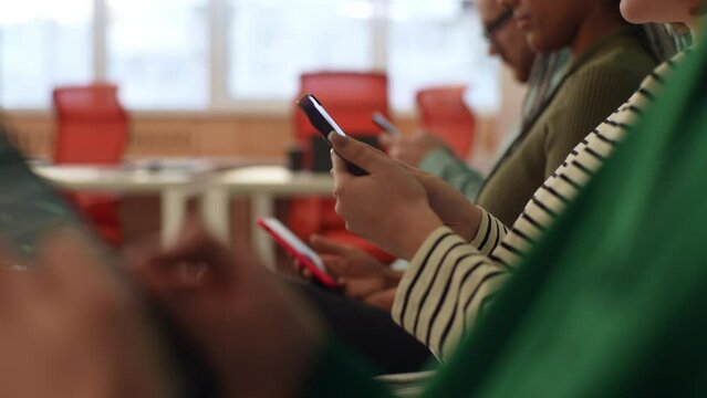 Closeup tracking shot of unrecognizable multicultural business people sitting in row using smartphones, waiting for job interview, sitting in queue line row in chairs in modern office waiting room.