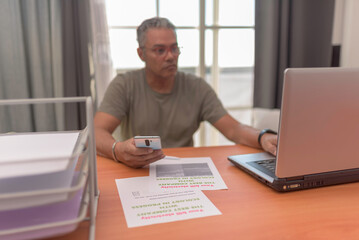 Mature man sits with laptop, paperwork, smartphone,addressing electricity bill dispute, he is...